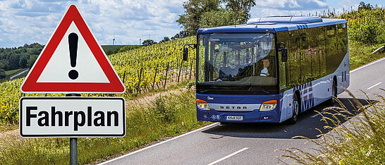 Auf dem Bild ist ein blauer Bus zu sehen, der auf einer Landstraße fährt. Rechts an der Straße steht ein Verkehrsschild mit der Aufschrift „Fahrplan“ und darüber ein Warnschild mit einem Ausrufezeichen, das zur Vorsicht mahnt
