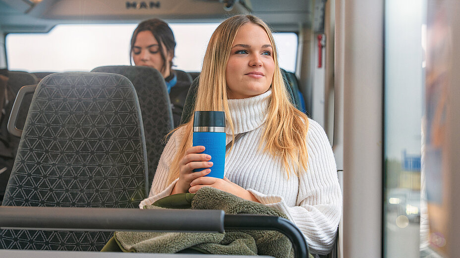 Eine sehr schöne junge blonde Frau in einem weißen Pullover sitzt am Fenster im Bus, blickt in die Ferne und hält einen blauen Thermobecher